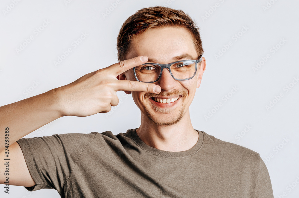 Young handsome man with glasses doing peace symbol with fingers over face, smiling cheerful showing victory. Handsome guy smiles over isolated gray background. Place for advertising.