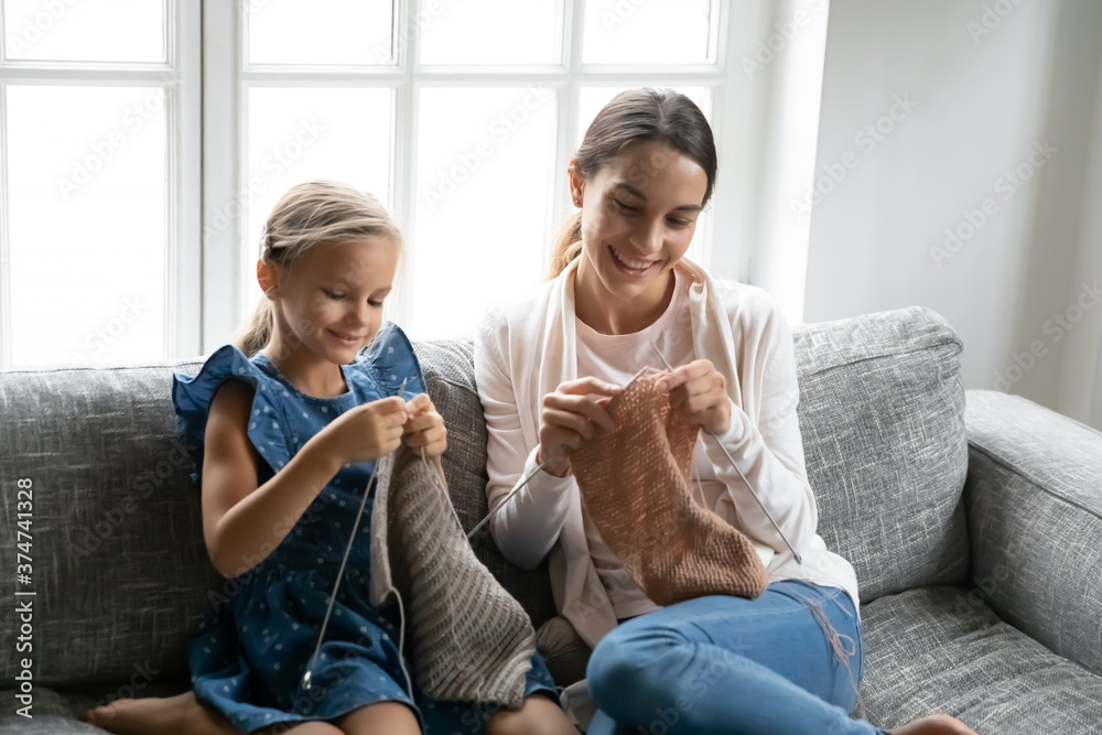 Φωτογραφία Smiling cute smart little kid girl resting on sofa with older sister or biracial nanny, involved in knitting warm clothes at home