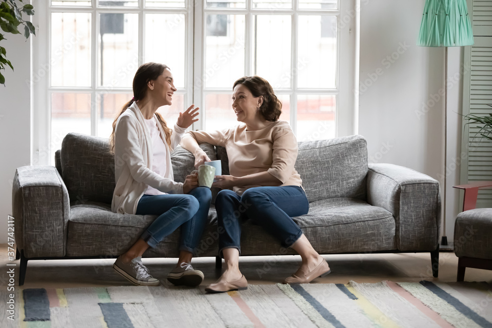 Full length joyful young mixed race woman drinking coffee with older 50s european mother, relaxing on comfortable couch. Happy elderly woman talking gossiping on weekend with grown daughter at home. Full length joyful young mixed race woman drinking coffee with older 50s european mother, relaxing on comfortable couch. Happy elderly woman talking gossiping on weekend with grown daughter at home.