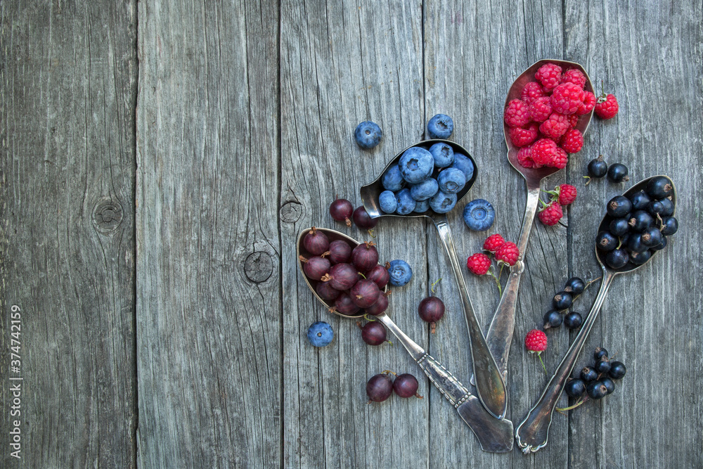 Gooseberries, black currants, raspberries and blueberries in spoons on a wooden old background.