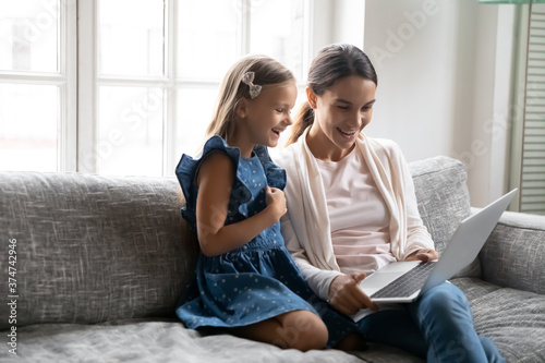 Happy little school age girl sitting on sofa with smiling mixed race nanny or mum, enjoying watching cartoons or comedian movie on laptop in living room, family weekend pastime hobby activity concept.