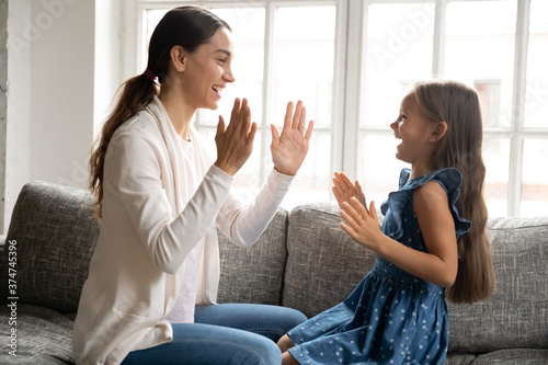 Smiling young mixed race woman playing patty cake game with laughing energetic small kid girl, sitting together on couch indoors. Happy multigenerational family enjoying weekend leisure time at home.