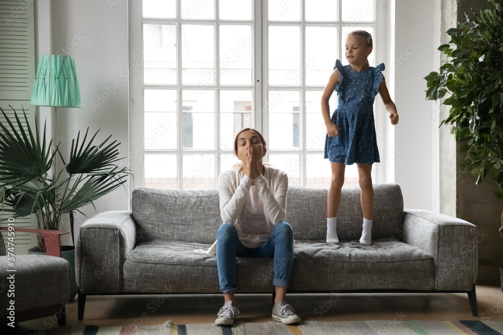 Φωτογραφία Full length energetic crazy little kid girl jumping on sofa near exhausted young biracial mother or babysitter