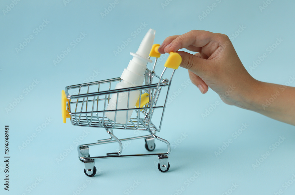 Nasal spray in white plastic packaging in a shopping cart, which is carried by the hand of a Caucasian man on a blue background. Selective focus. Purchase at a pharmacy