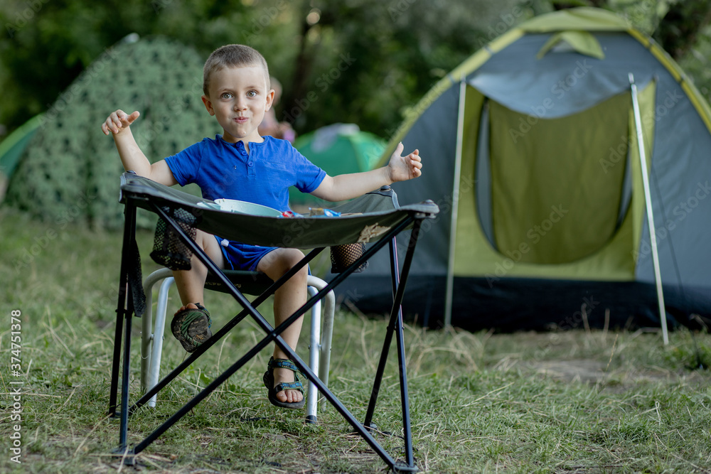 Little boy eating breakfast outdoor at camping site in nature sunlight with a tent at the background. Little boy eating breakfast outdoor at camping site in nature sunlight with a tent at the background.