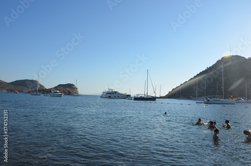 A land view of yachts, sailboats and people swimming on the shore in a bay in the Aegean Sea. Aegean islands visible behind sailboats and yachts. People swimming and having fun on the shore side. 