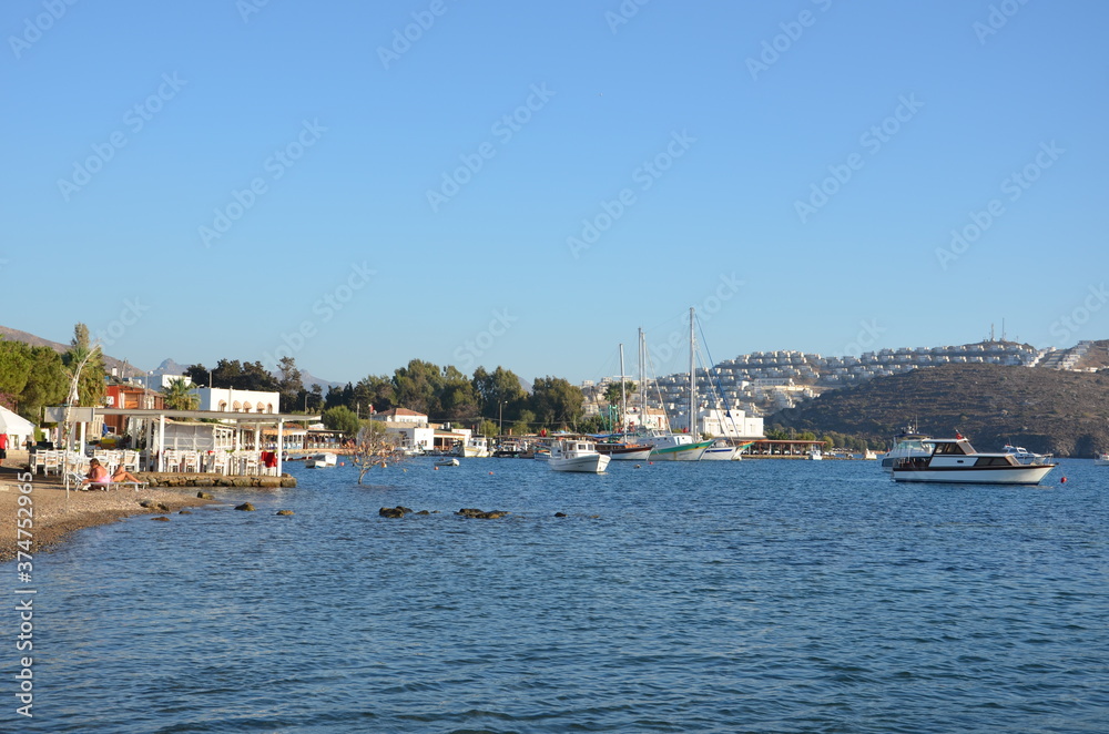 View of sailboats, boats and Aegean islands from the beach. Sunlight reflected on the sea surface. Settlements and restaurants on the opposite shore. Side view of the entire bay.
