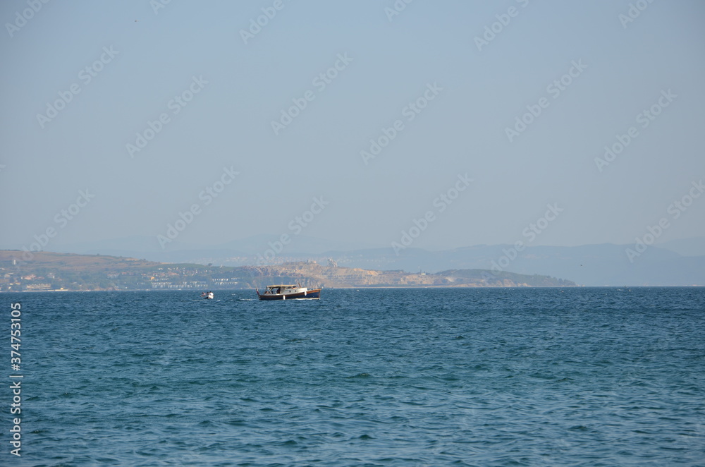 A small fishing boat and a small dinghy in the immense Aegean Sea. A background with a hazy view of distant hills and residential areas. The hills on the opposite shore appear in silhouette. 