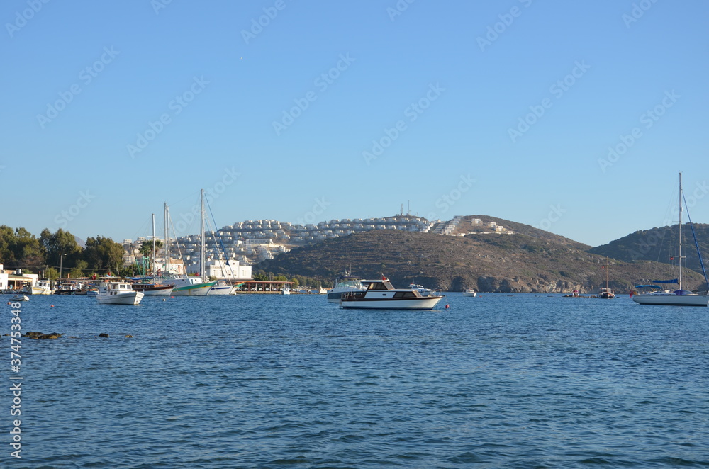 View of sailboats, boats and Aegean islands from the beach. Sunlight reflected on the sea surface. Settlements and restaurants on the opposite shore. Side view of the entire bay.