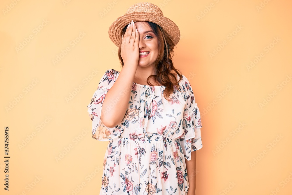Young beautiful caucasian woman wearing summer dress and hat covering one eye with hand, confident smile on face and surprise emotion. Young beautiful caucasian woman wearing summer dress and hat covering one eye with hand, confident smile on face and surprise emotion.