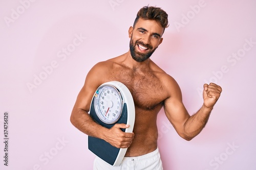 Young hispanic man standing shirtless holding weighing machine screaming proud  celebrating victory and success very excited with raised arms