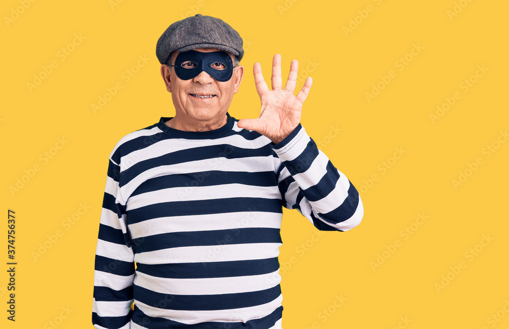Senior handsome man wearing burglar mask and t-shirt showing and pointing up with fingers number five while smiling confident and happy. Senior handsome man wearing burglar mask and t-shirt showing and pointing up with fingers number five while smiling confident and happy.