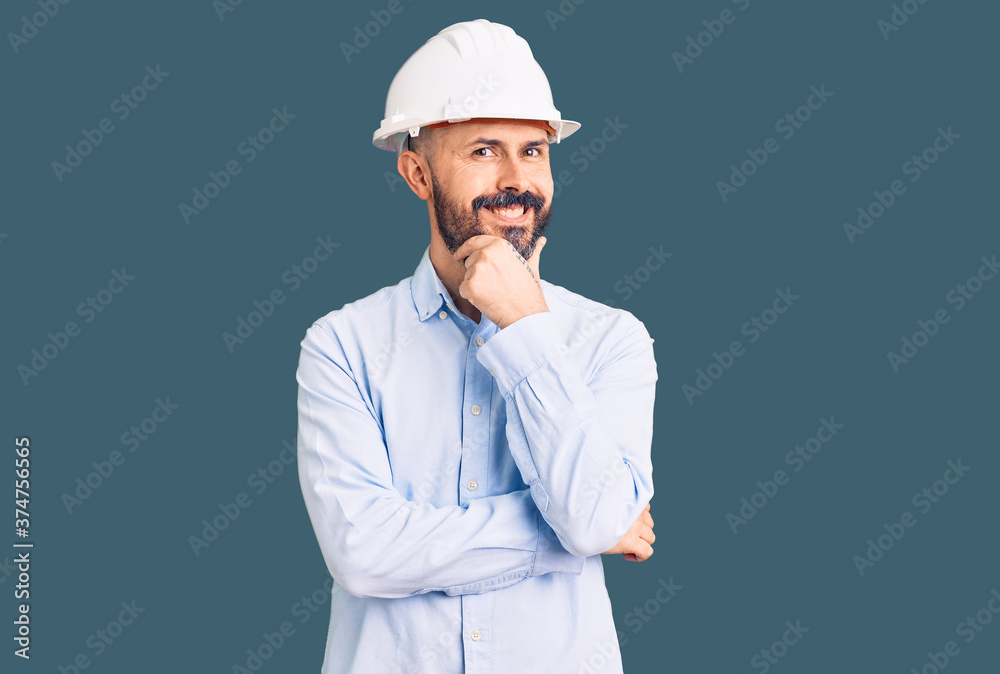 Young handsome man wearing architect hardhat looking confident at the camera smiling with crossed arms and hand raised on chin. thinking positive. Young handsome man wearing architect hardhat looking confident at the camera smiling with crossed arms and hand raised on chin. thinking positive.