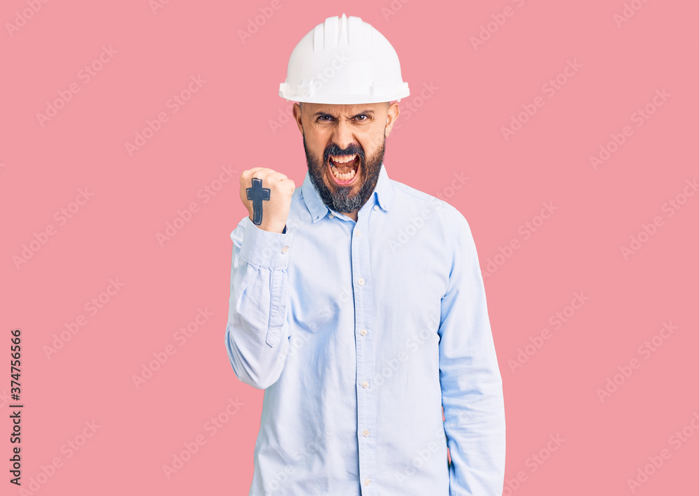 Young handsome man wearing architect hardhat angry and mad raising fist frustrated and furious while shouting with anger. rage and aggressive concept. Young handsome man wearing architect hardhat angry and mad raising fist frustrated and furious while shouting with anger. rage and aggressive concept.
