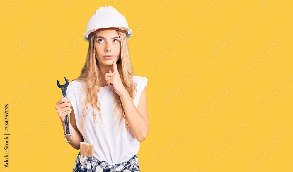 Beautiful caucasian woman with blonde hair wearing hardhat and builder clothes serious face thinking about question with hand on chin, thoughtful about confusing idea