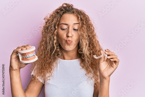 Beautiful caucasian teenager girl holding invisible aligner orthodontic and braces making fish face with mouth and squinting eyes, crazy and comical.