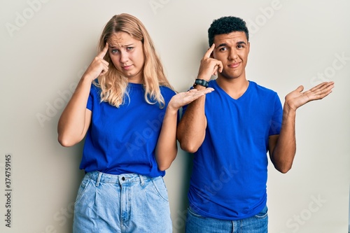 Young interracial couple wearing casual clothes confused and annoyed with open palm showing copy space and pointing finger to forehead. think about it.