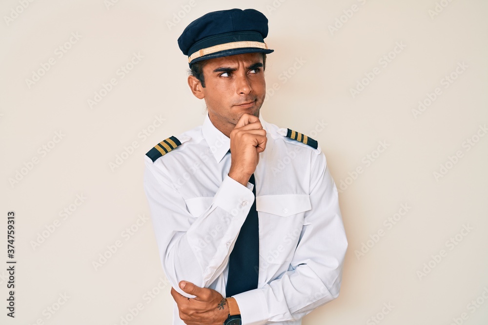 Young hispanic man wearing airplane pilot uniform with hand on chin thinking about question, pensive expression. smiling with thoughtful face. doubt concept. Young hispanic man wearing airplane pilot uniform with hand on chin thinking about question, pensive expression. smiling with thoughtful face. doubt concept.