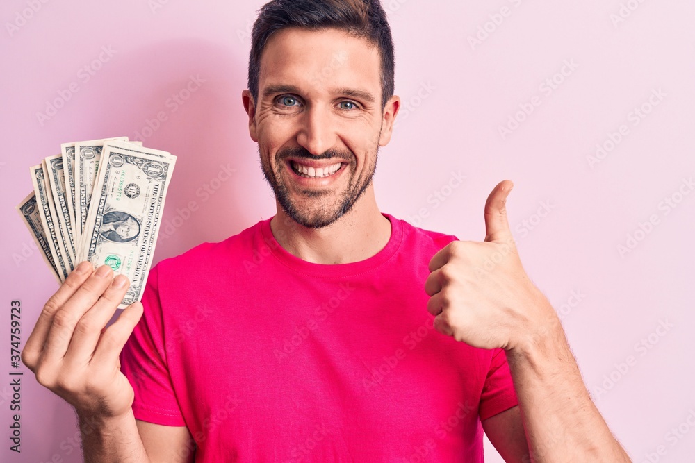 Young handsome man holding bunch of dollars banknotes over isolated pink background smiling happy and positive, thumb up doing excellent and approval sign Young handsome man holding bunch of dollars banknotes over isolated pink background smiling happy and positive, thumb up doing excellent and approval sign