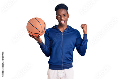 Young african american man holding basketball ball pointing thumb up to the side smiling happy with open mouth