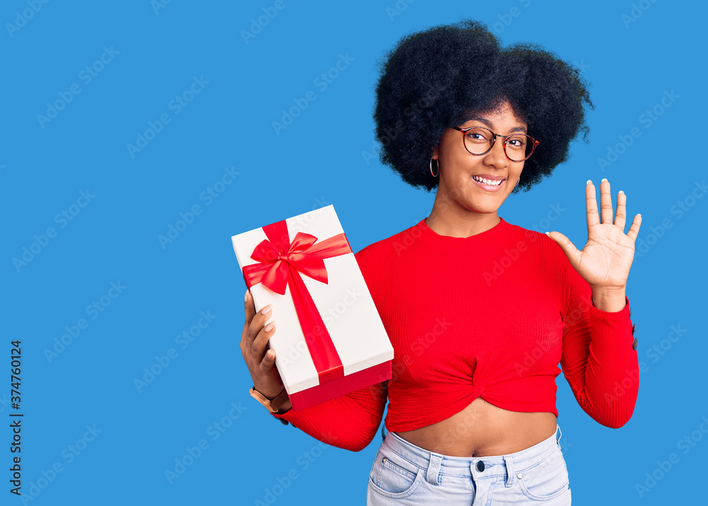 Young african american girl holding gift waiving saying hello happy and smiling, friendly welcome gesture Young african american girl holding gift waiving saying hello happy and smiling, friendly welcome gesture
