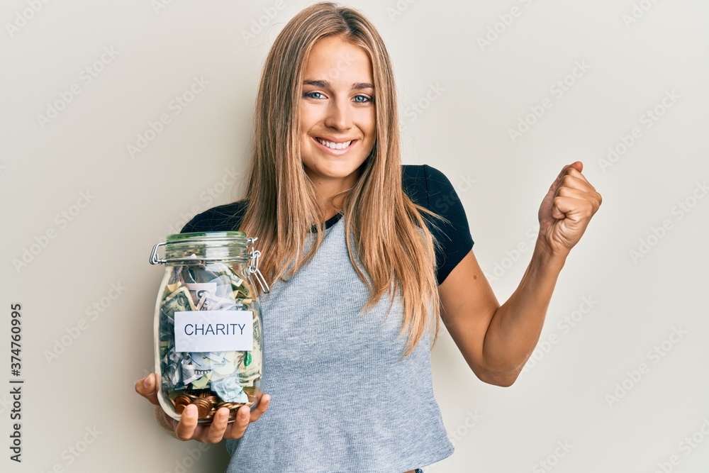 Obraz premium Young blonde woman holding charity jar with money screaming proud, celebrating victory and success very excited with raised arm