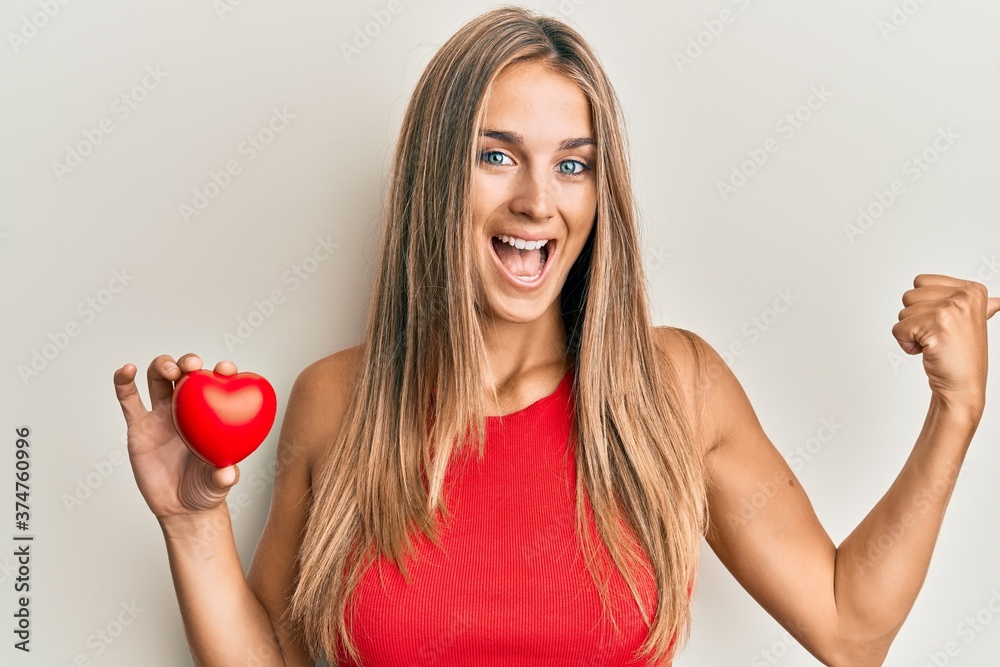Young blonde woman holding heart pointing thumb up to the side smiling happy with open mouth Young blonde woman holding heart pointing thumb up to the side smiling happy with open mouth