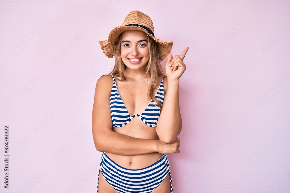 Young beautiful blonde woman wearing bikini and hat smiling happy pointing with hand and finger to the side Young beautiful blonde woman wearing bikini and hat smiling happy pointing with hand and finger to the side