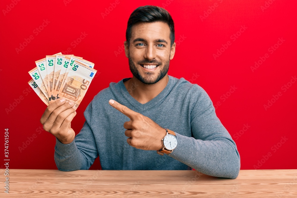 Handsome hispanic man holding bunch of 50 euro banknotes smiling happy pointing with hand and finger Handsome hispanic man holding bunch of 50 euro banknotes smiling happy pointing with hand and finger