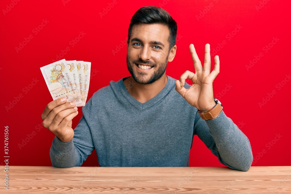 Handsome hispanic man holding united kingdom pounds doing ok sign with fingers, smiling friendly gesturing excellent symbol Handsome hispanic man holding united kingdom pounds doing ok sign with fingers, smiling friendly gesturing excellent symbol