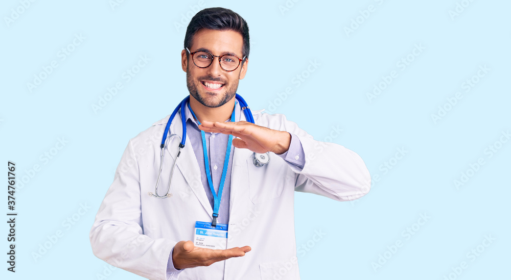 Young hispanic man wearing doctor uniform and stethoscope gesturing with hands showing big and large size sign, measure symbol. smiling looking at the camera. measuring concept. Young hispanic man wearing doctor uniform and stethoscope gesturing with hands showing big and large size sign, measure symbol. smiling looking at the camera. measuring concept.