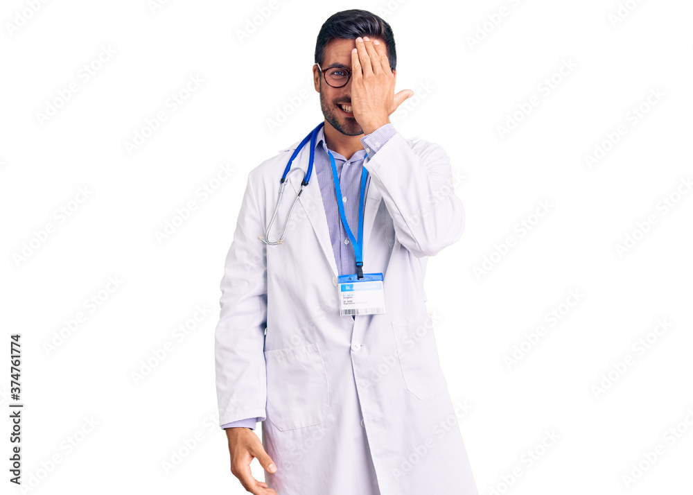 Young hispanic man wearing doctor uniform and stethoscope covering one eye with hand, confident smile on face and surprise emotion.