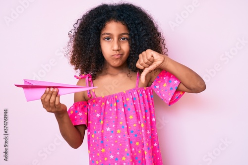 African american child with curly hair holding paper airplane with angry face, negative sign showing dislike with thumbs down, rejection concept