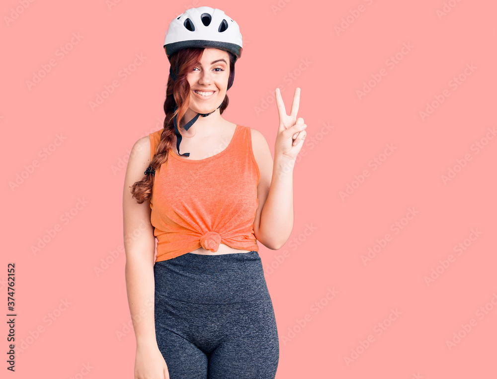 Young beautiful woman wearing bike helmet smiling with happy face winking at the camera doing victory sign. number two.