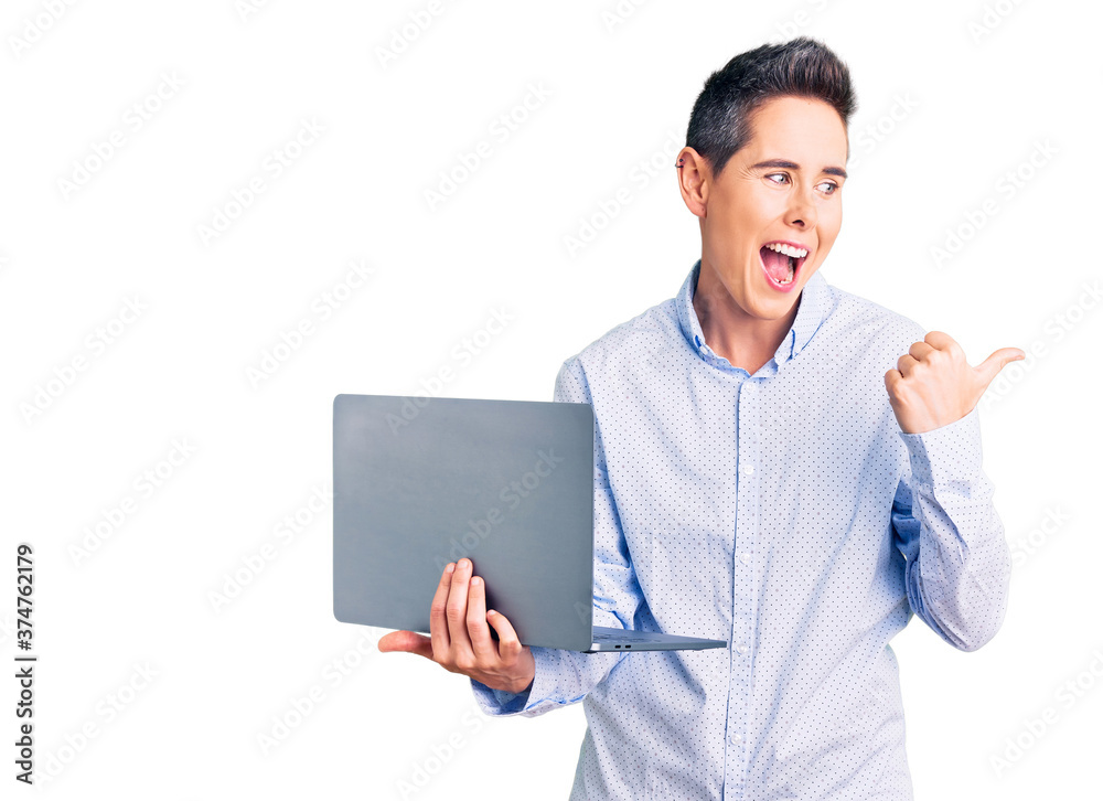 Young woman with short hair holding laptop pointing thumb up to the side smiling happy with open mouth Young woman with short hair holding laptop pointing thumb up to the side smiling happy with open mouth