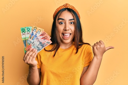 Young latin woman holding australian dollars banknotes pointing thumb up to the side smiling happy with open mouth