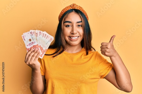 Young latin woman holding colombian pesos smiling happy and positive, thumb up doing excellent and approval sign