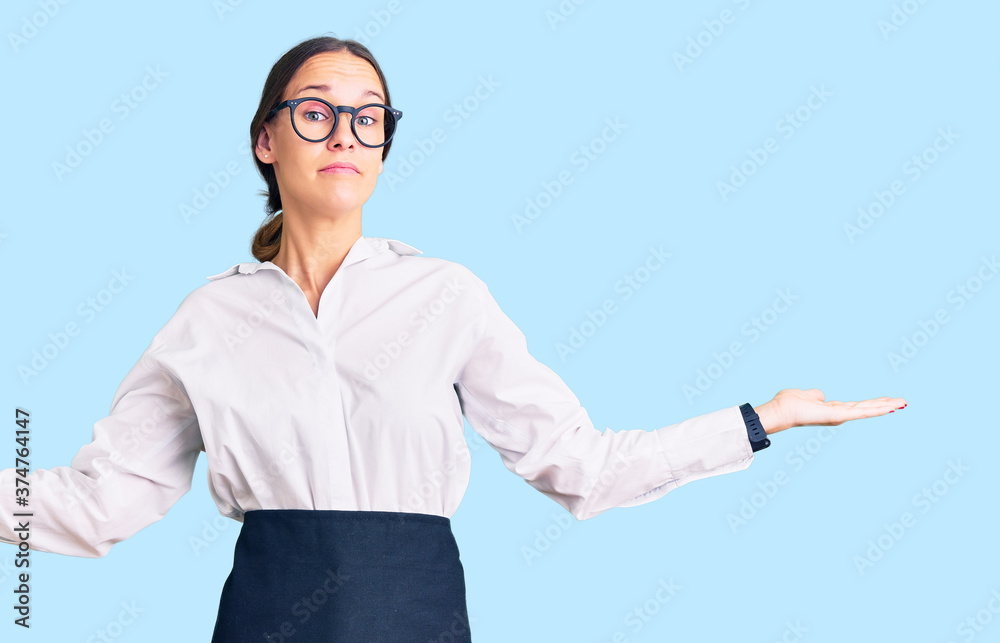 Beautiful brunette young woman wearing professional waitress apron clueless and confused expression with arms and hands raised. doubt concept. Beautiful brunette young woman wearing professional waitress apron clueless and confused expression with arms and hands raised. doubt concept.