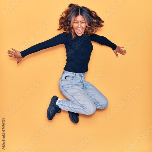 Young beautiful latin woman smiling happy. Jumping with smile on face and arms opened over isolated yellow background