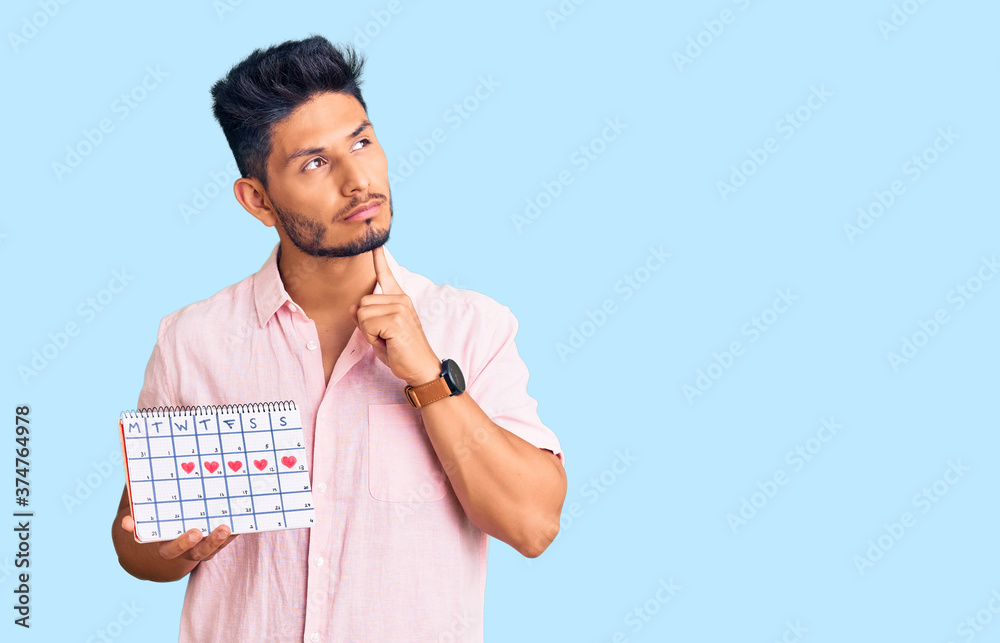 Handsome latin american young man holding heart calendar serious face thinking about question with hand on chin, thoughtful about confusing idea Handsome latin american young man holding heart calendar serious face thinking about question with hand on chin, thoughtful about confusing idea