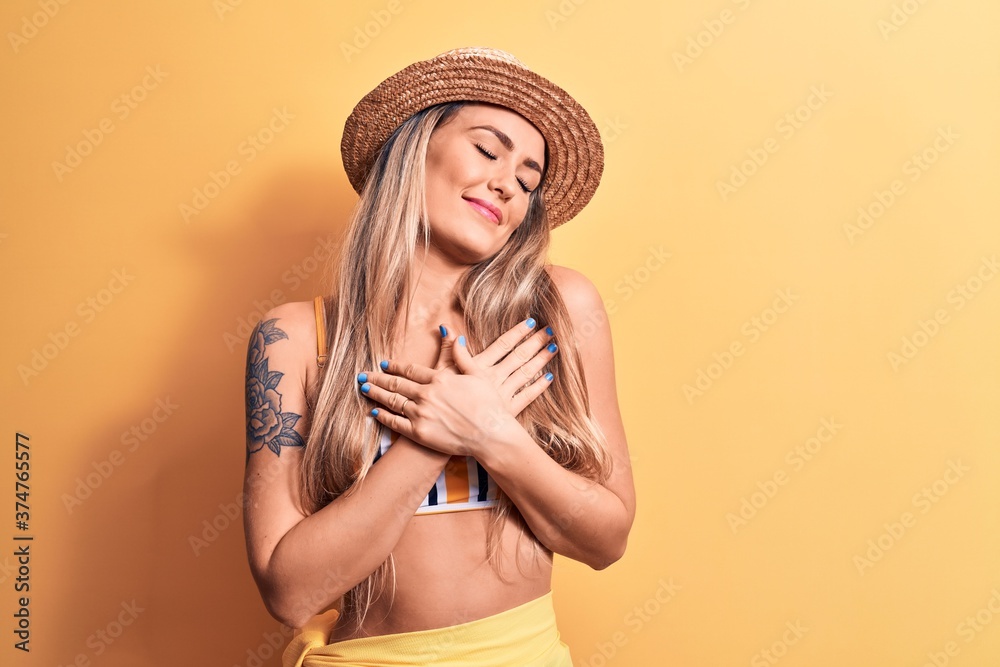 Young beautiful blonde woman wearing bikini and summer hat over isolated yellow background smiling with hands on chest, eyes closed with grateful gesture on face. Health concept. Young beautiful blonde woman wearing bikini and summer hat over isolated yellow background smiling with hands on chest, eyes closed with grateful gesture on face. Health concept.