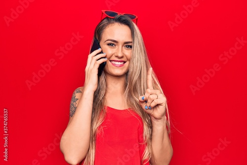 Beautiful blonde woman having conversation talking on the smartphone over red background smiling with an idea or question pointing finger with happy face, number one