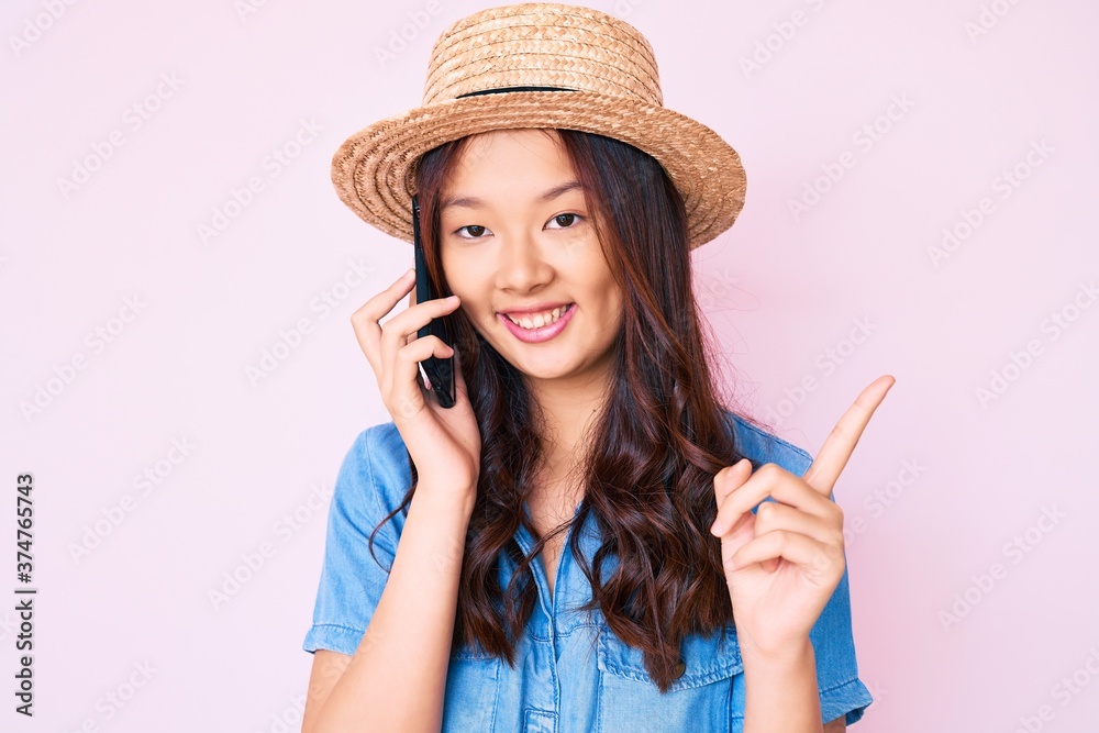 Young beautiful chinese girl wearing summer hat having conversation talking on the smartphone smiling happy pointing with hand and finger to the side