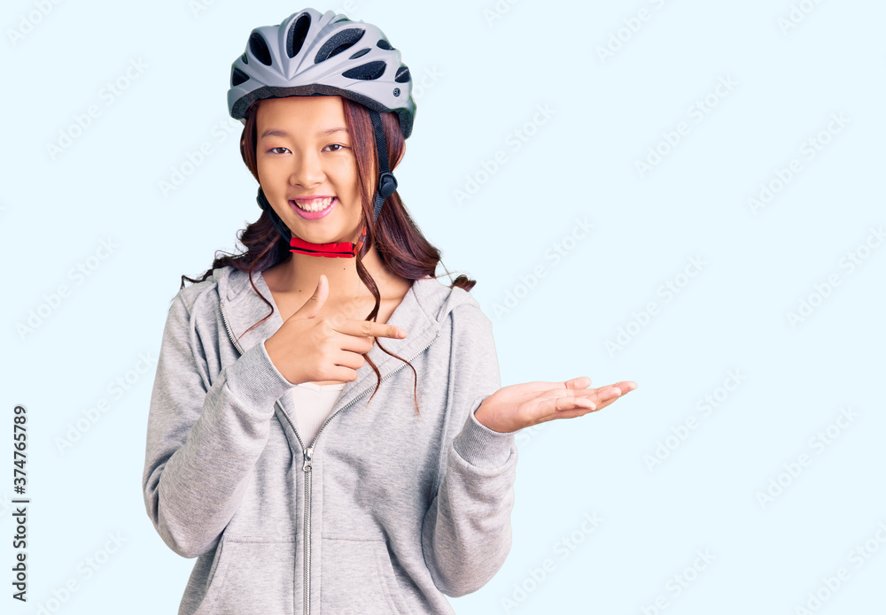 Young beautiful chinese girl wearing bike helmet amazed and smiling to the camera while presenting with hand and pointing with finger. Young beautiful chinese girl wearing bike helmet amazed and smiling to the camera while presenting with hand and pointing with finger.
