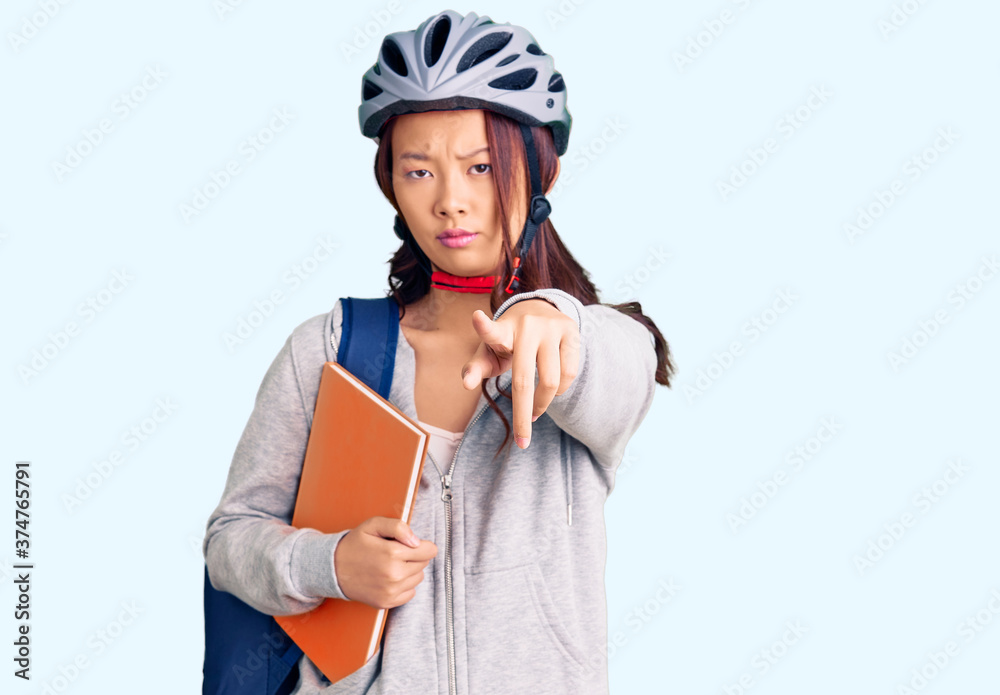 Young beautiful chinese girl wearing student backpack and bike helmet holding book pointing with finger to the camera and to you, confident gesture looking serious Young beautiful chinese girl wearing student backpack and bike helmet holding book pointing with finger to the camera and to you, confident gesture looking serious