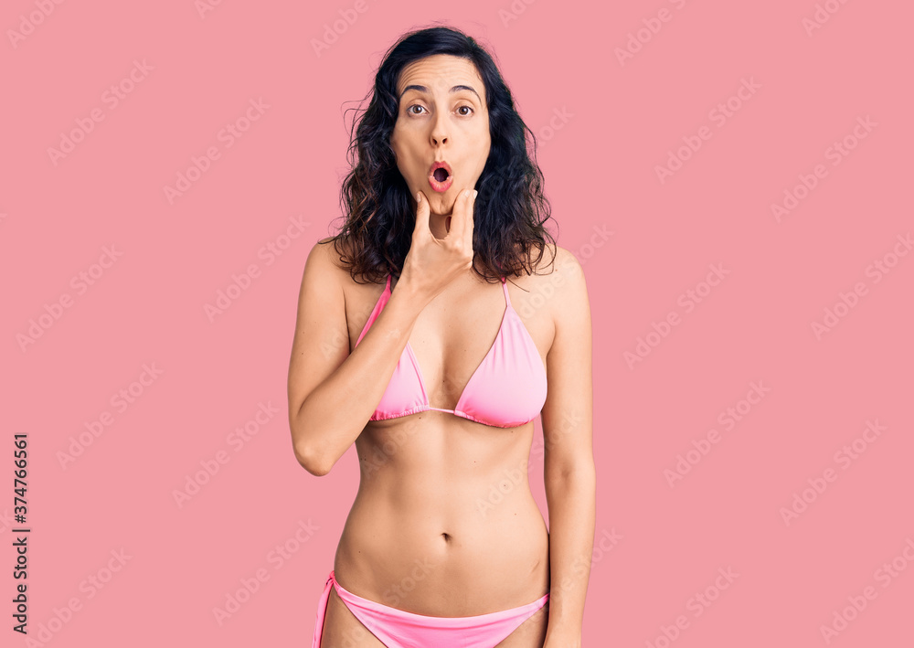 Young beautiful hispanic woman wearing bikini looking fascinated with disbelief, surprise and amazed expression with hands on chin Young beautiful hispanic woman wearing bikini looking fascinated with disbelief, surprise and amazed expression with hands on chin