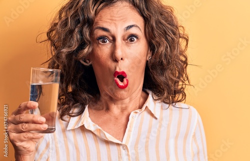 Middle age brunette woman drinking glass of water standing over isolated yellow background scared and amazed with open mouth for surprise, disbelief face © Krakenimages.com
