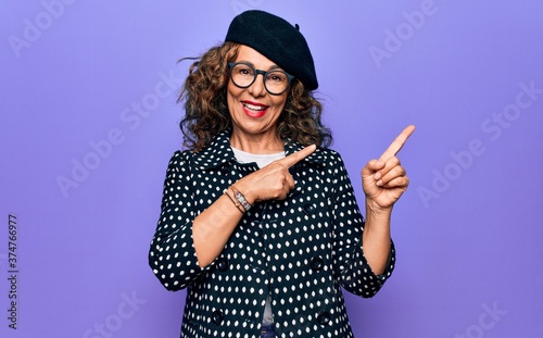 Middle age beautiful woman wearing casual jacket and french beret over purple background smiling and looking at the camera pointing with two hands and fingers to the side.