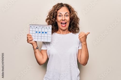 Middle age beautiful woman holding climatology calendar showing cloudy and rainy weather pointing thumb up to the side smiling happy with open mouth