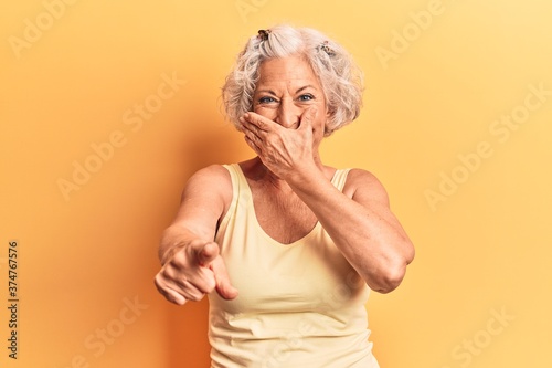 Senior grey-haired woman wearing casual clothes laughing at you, pointing finger to the camera with hand over mouth, shame expression